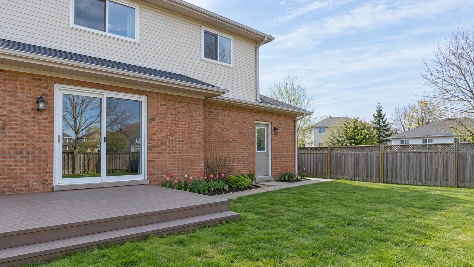 Back of a two-storey 1990s brick and vinyl house, a white sliding patio door opening onto a low deck and a grey side door with a small glass lite beside it